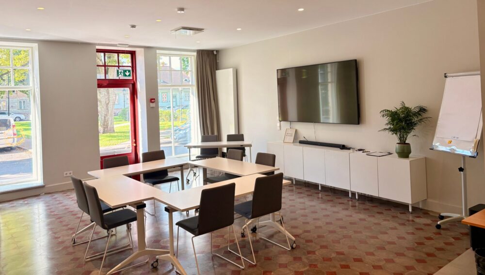 Valgusküllane Park Hotell Viljandi seminariruum U-kujuliselt paigutatud laudade, hallide toolide, seinale kinnitatud suure teleri, pabertahvli ning parki avanevate kõrgete akendega.